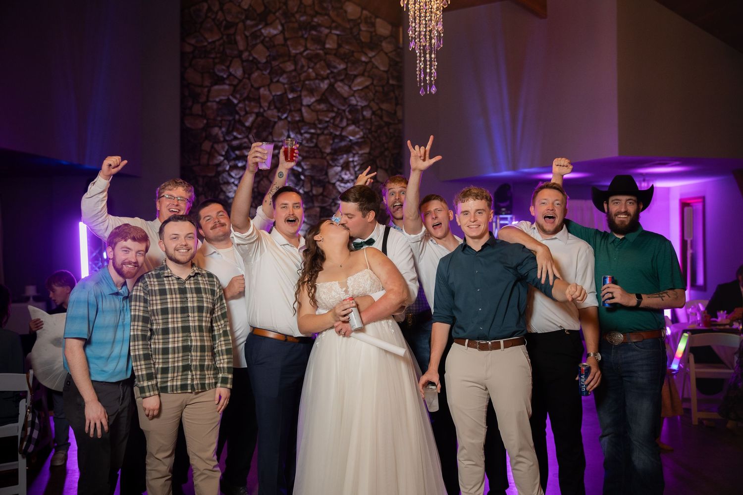 Bride in white gown poses with groomsmen celebrating at wedding reception with purple lighting.