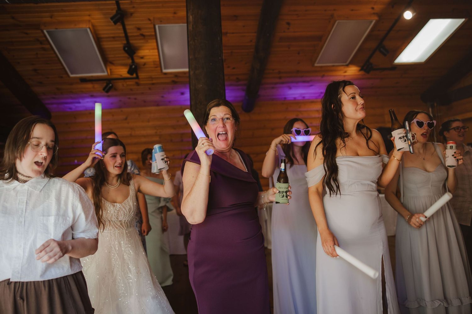 Bridesmaids and guests dance with glow sticks under purple lights at festive wedding reception.