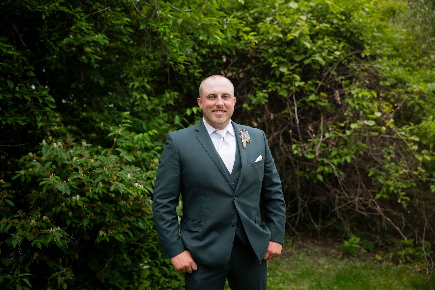 Groom in dark green three-piece suit with boutonniere stands confidently in front of green foliage outdoors.