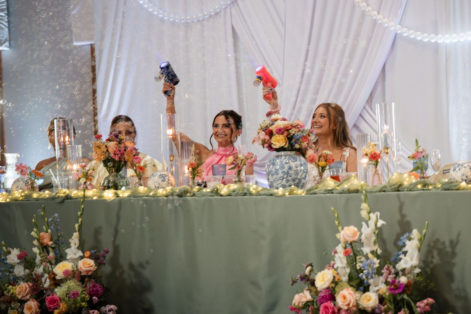 Bridesmaids raise colorful party favors at floral-decorated head table during wedding reception celebration.