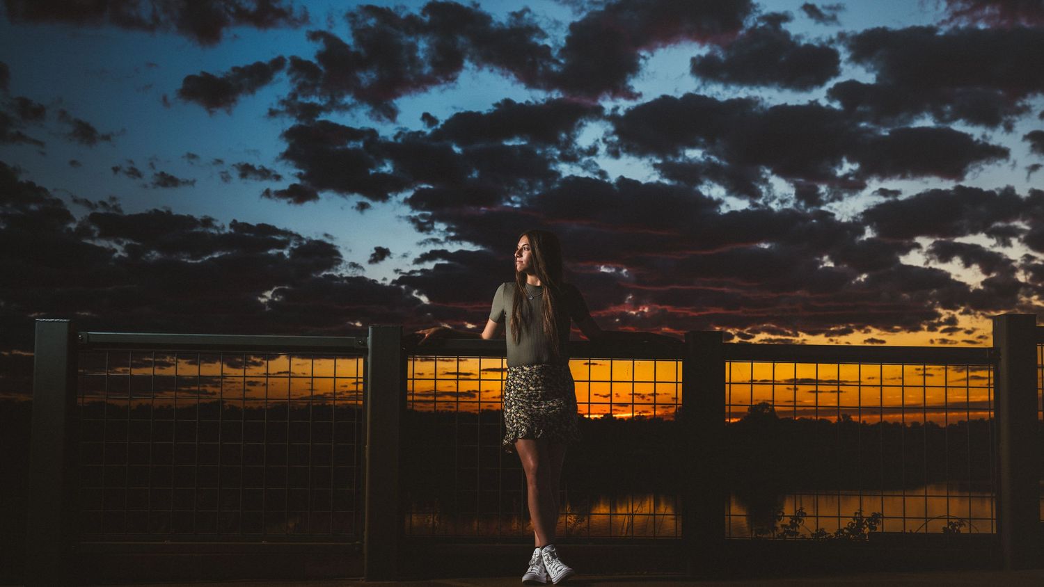 A young woman stands by a fence at sunset with dramatic dark clouds filling the twilight sky behind her.