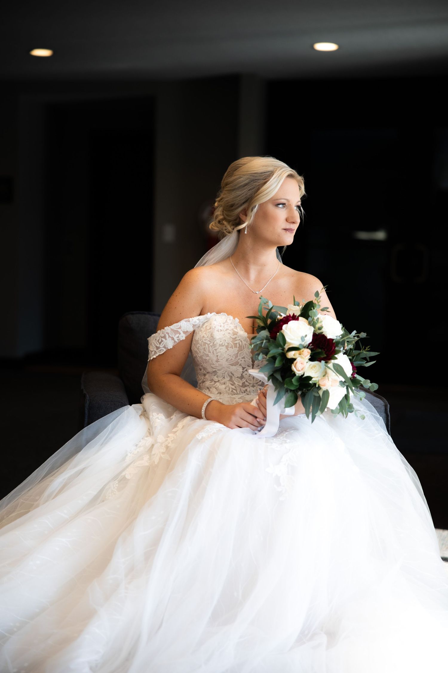 Bride in off-shoulder lace ballgown holding red and white bouquet, seated and gazing thoughtfully to the side.