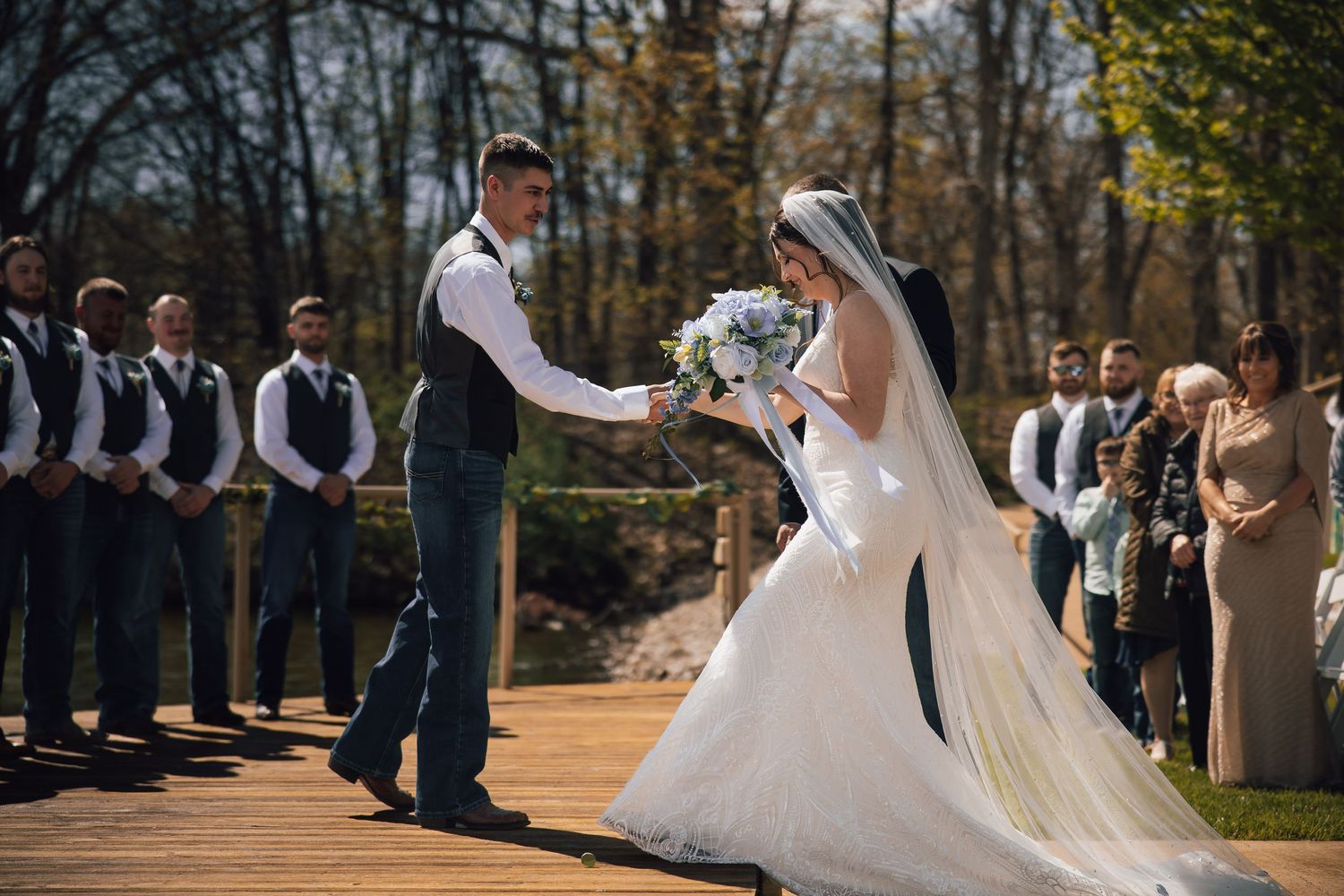 Bride and groom hold hands during outdoor wedding ceremony on wooden deck with groomsmen and guests watching.