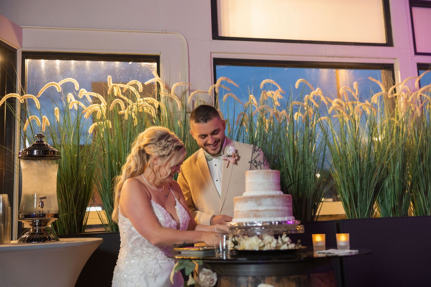 Couple cuts their white wedding cake together in front of decorative grass wall display.