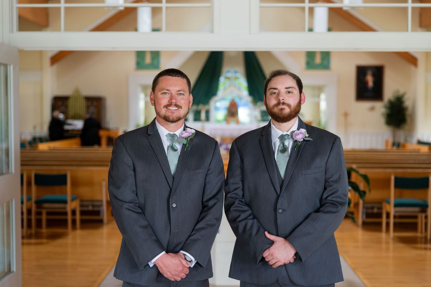 Two groomsmen in gray suits with mint ties stand together inside a church before a wedding ceremony.