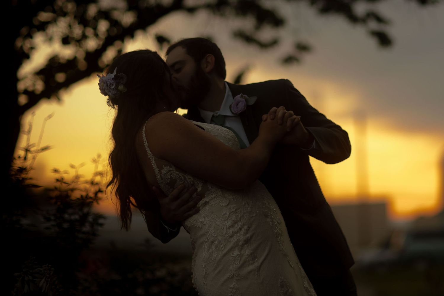 A bride and groom share a romantic kiss at sunset, silhouetted against a warm golden sky.