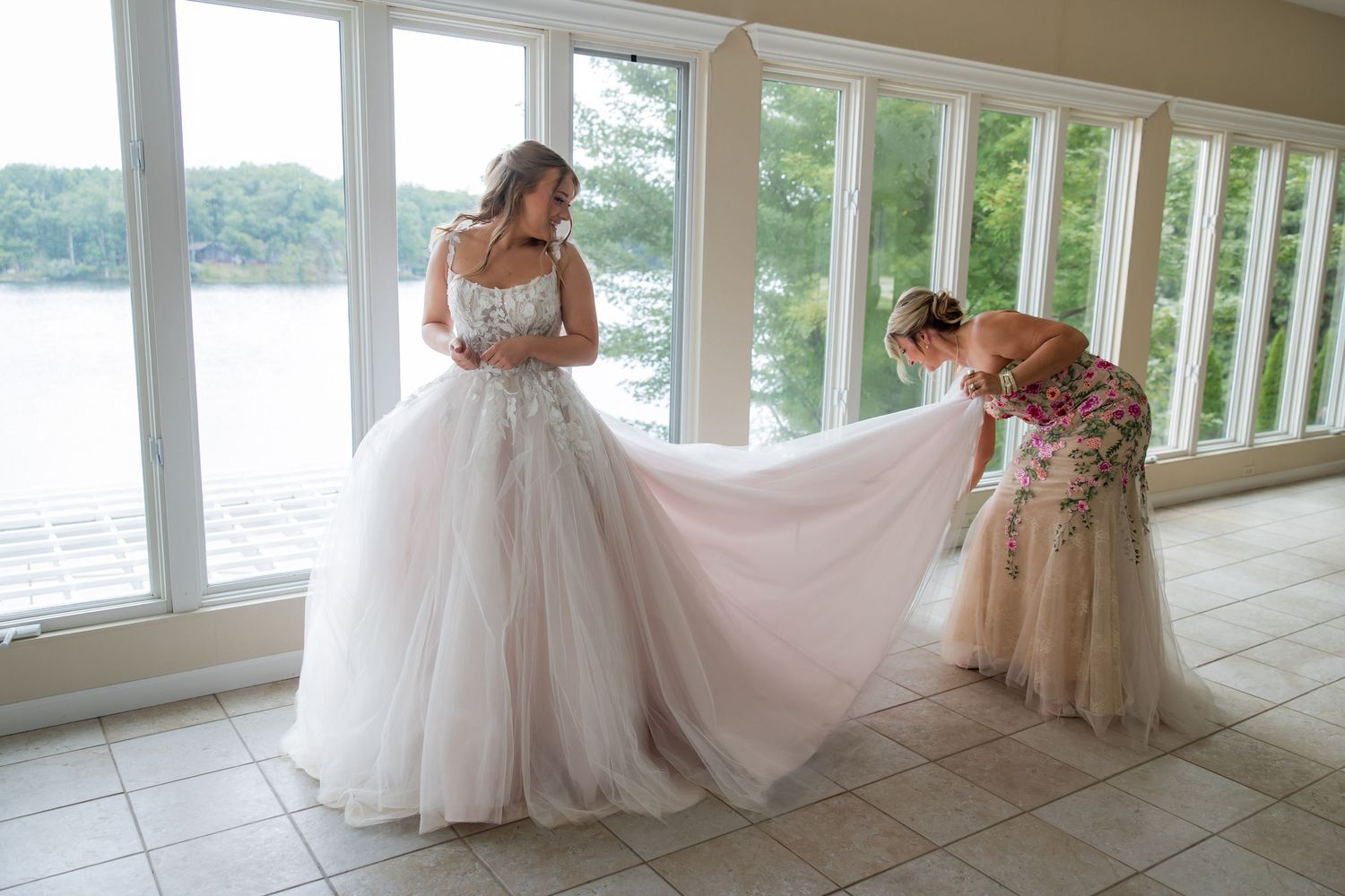 Bride in elegant white tulle ball gown being helped with her dress train near large windows overlooking a lake.