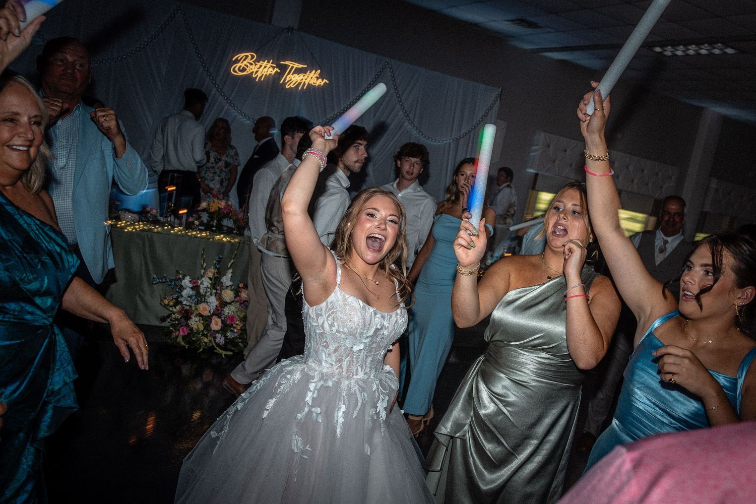 Excited bride in white gown celebrates with bridesmaids holding glowing light sticks on a lively wedding dance floor.