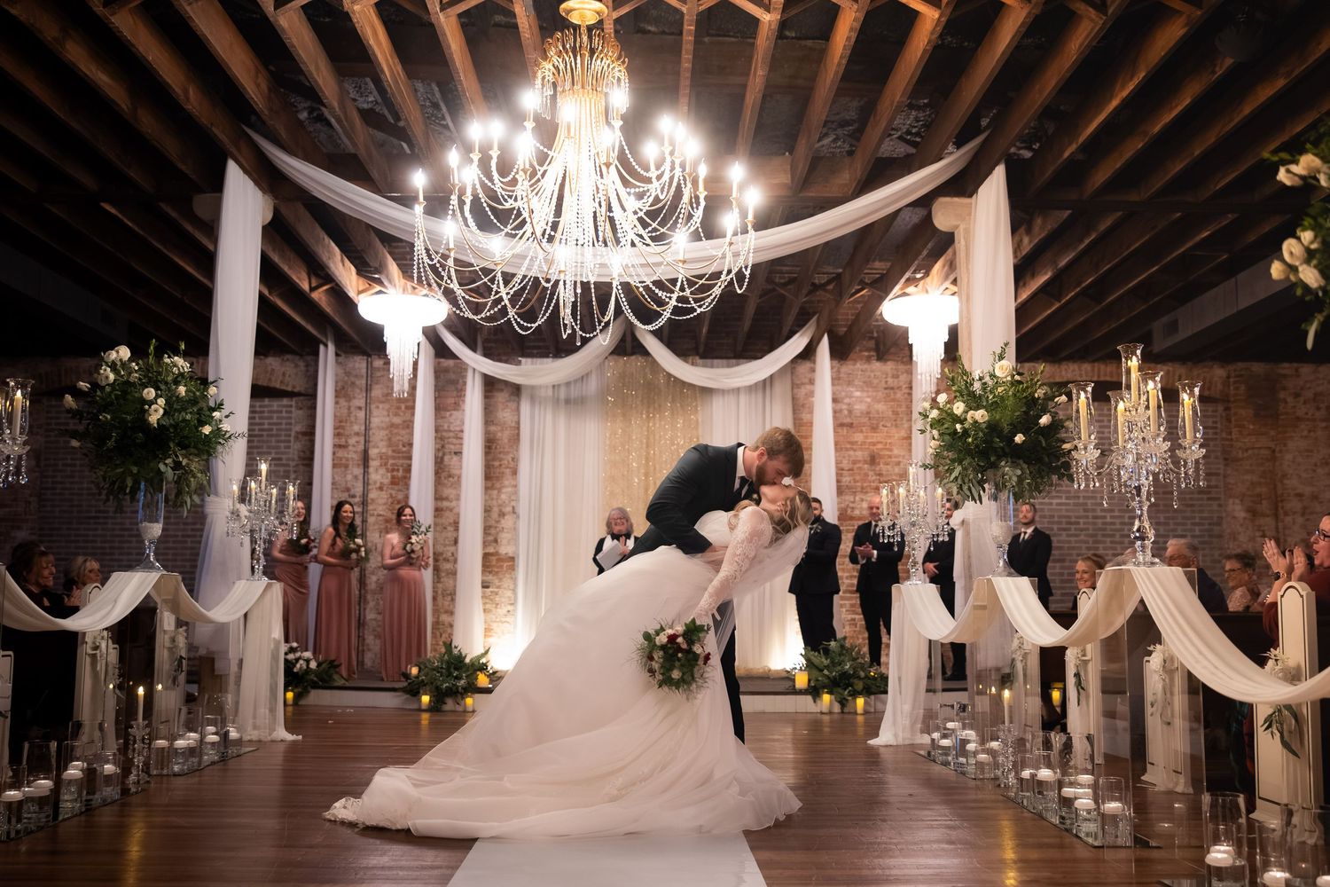 Groom dips bride in elegant wedding venue with chandelier, draped fabric, and floral arrangements during first dance.