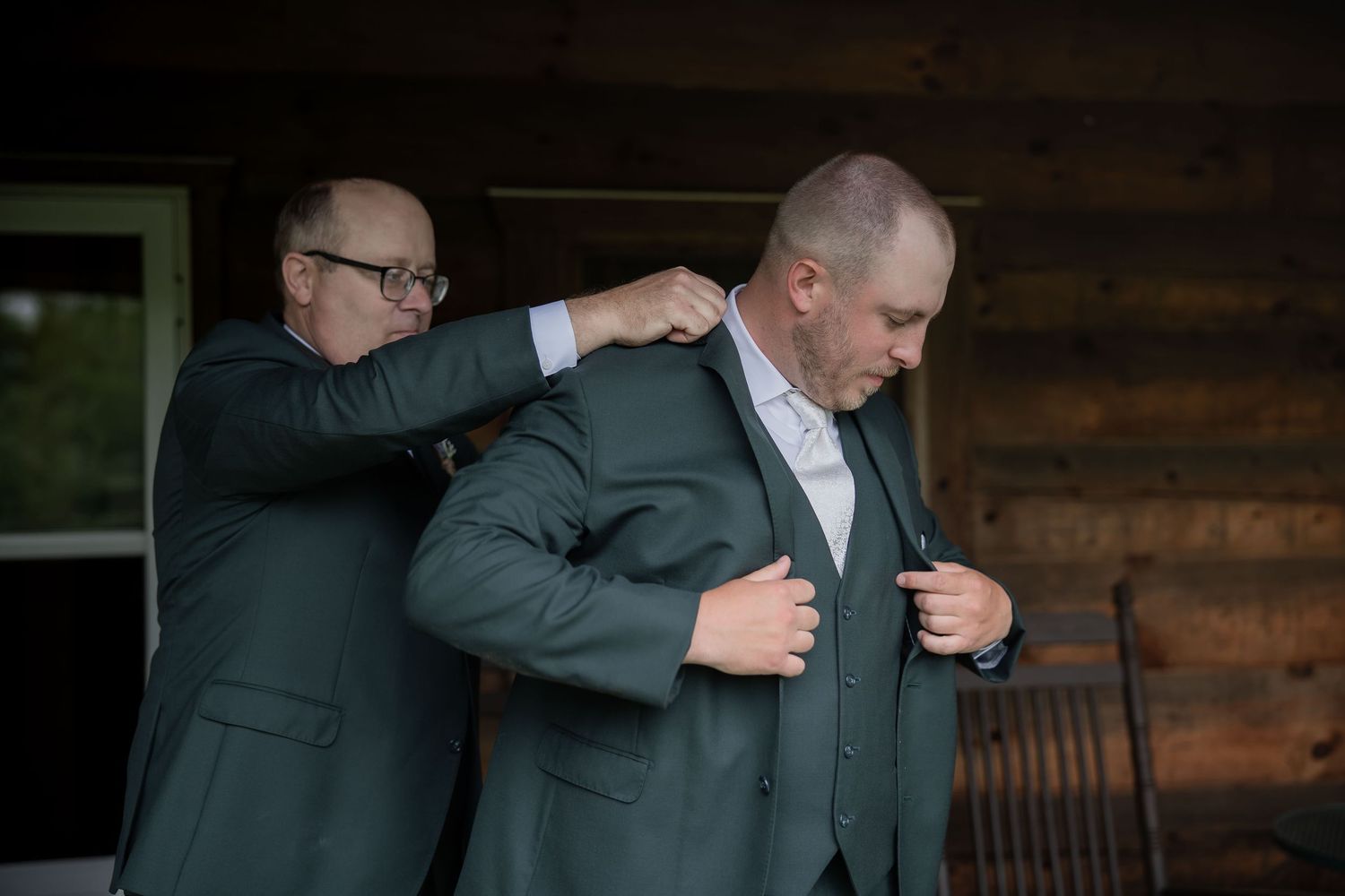 Two men in gray suits with one helping the other adjust his jacket outside a rustic log cabin.