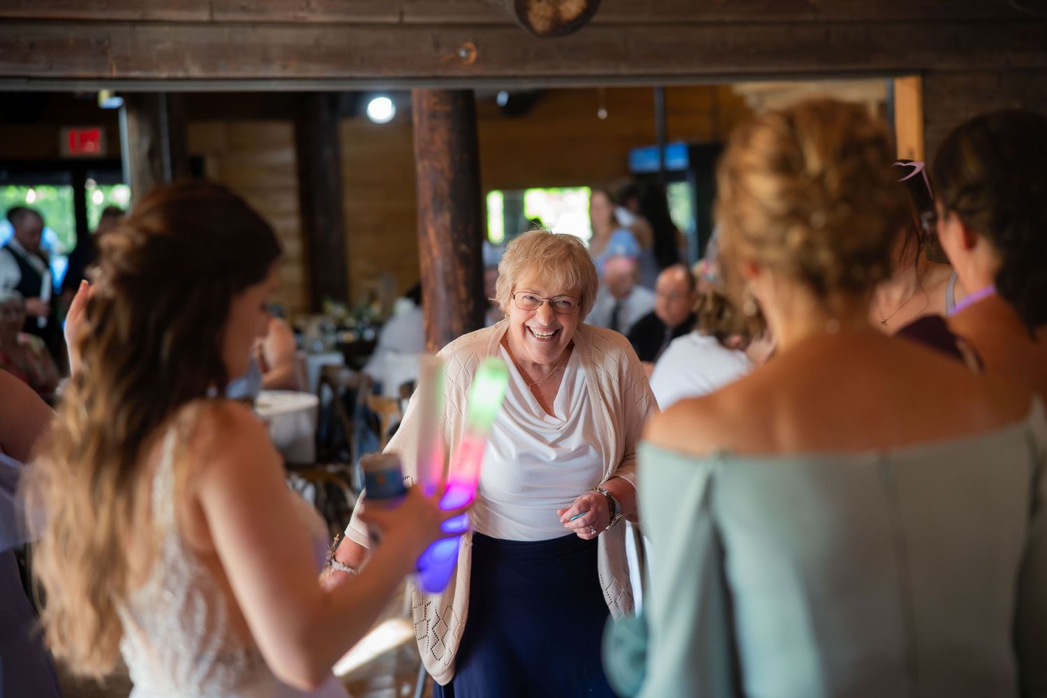 Joyful elderly woman dances and smiles with guests at a warmly lit wedding reception.