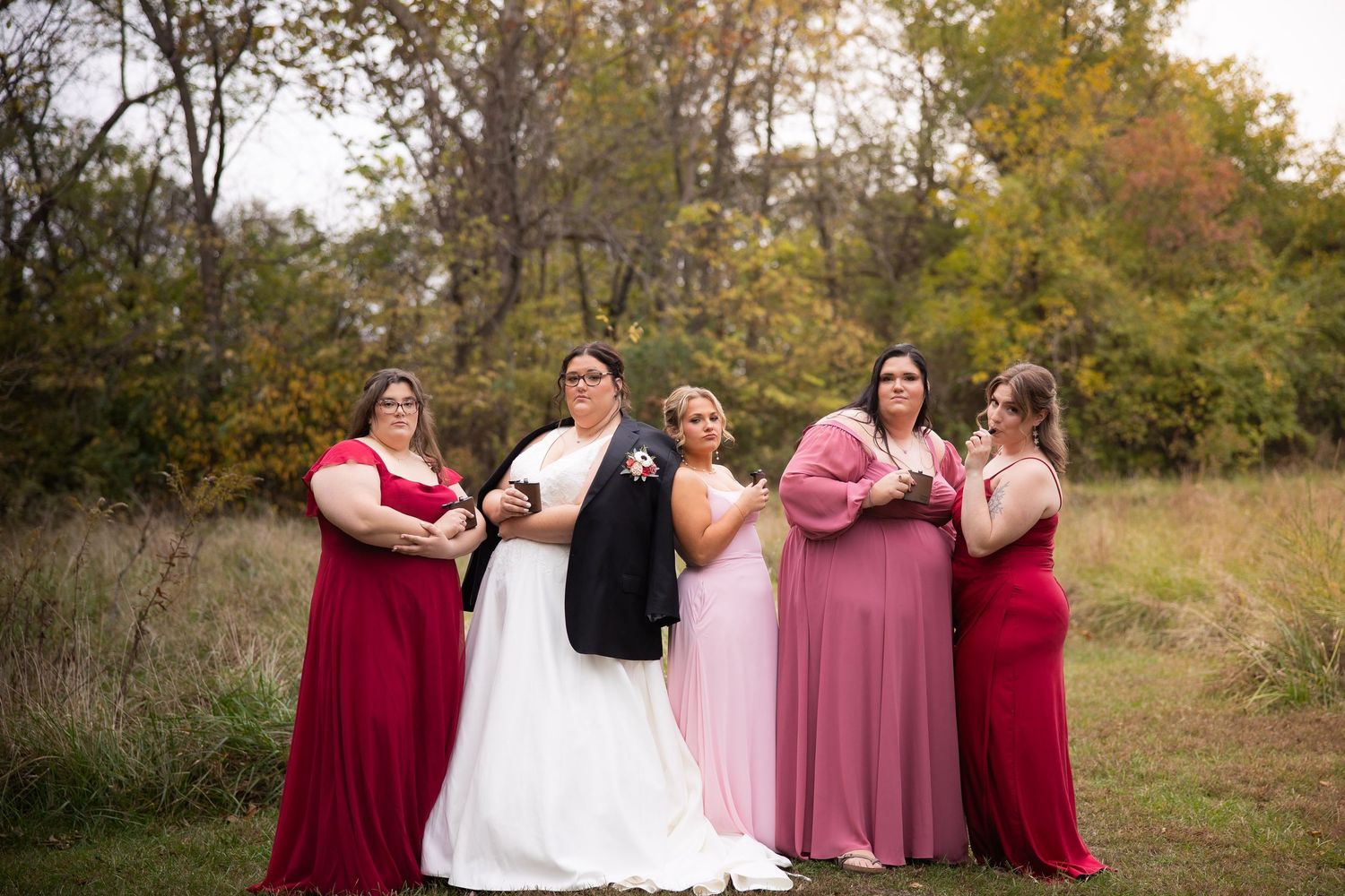 Wedding party poses in burgundy and pink dresses against a scenic fall backdrop with trees.
