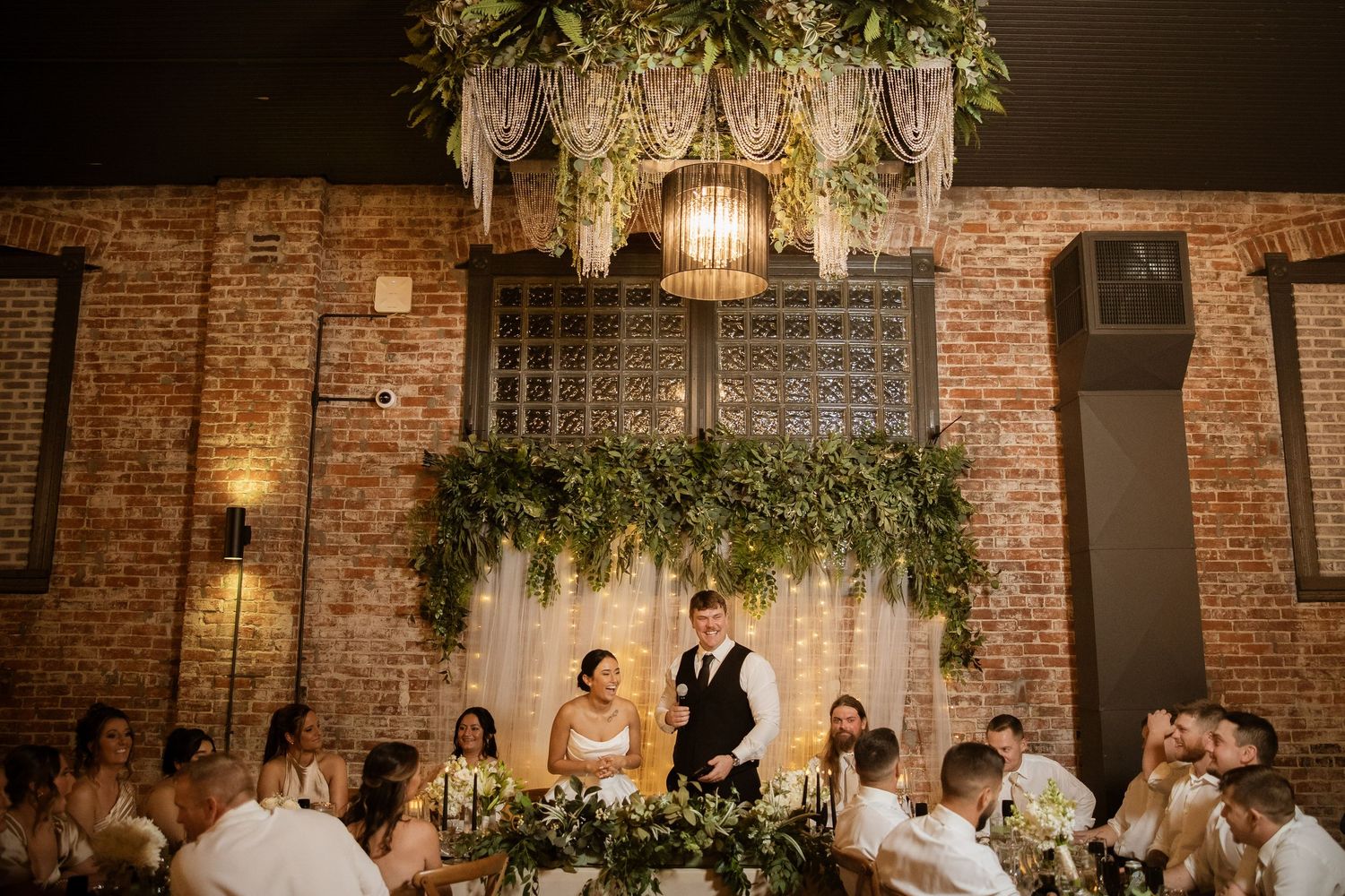 Wedding ceremony setup in rustic brick venue with greenery and floral decorations illuminated by warm lighting.
