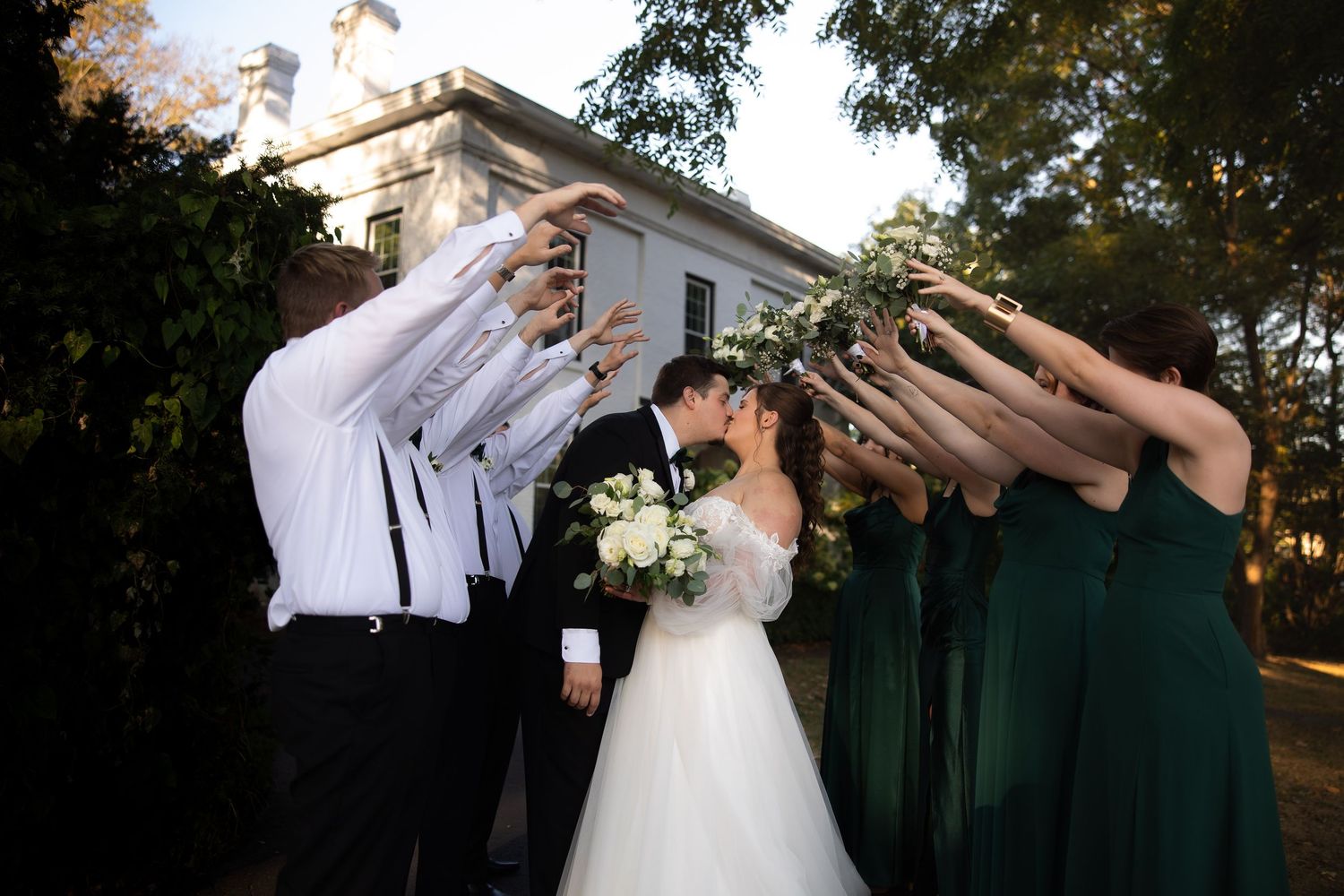 Bride and groom kissing under an arch of raised hands formed by wedding party in green dresses and black suits outdoors.