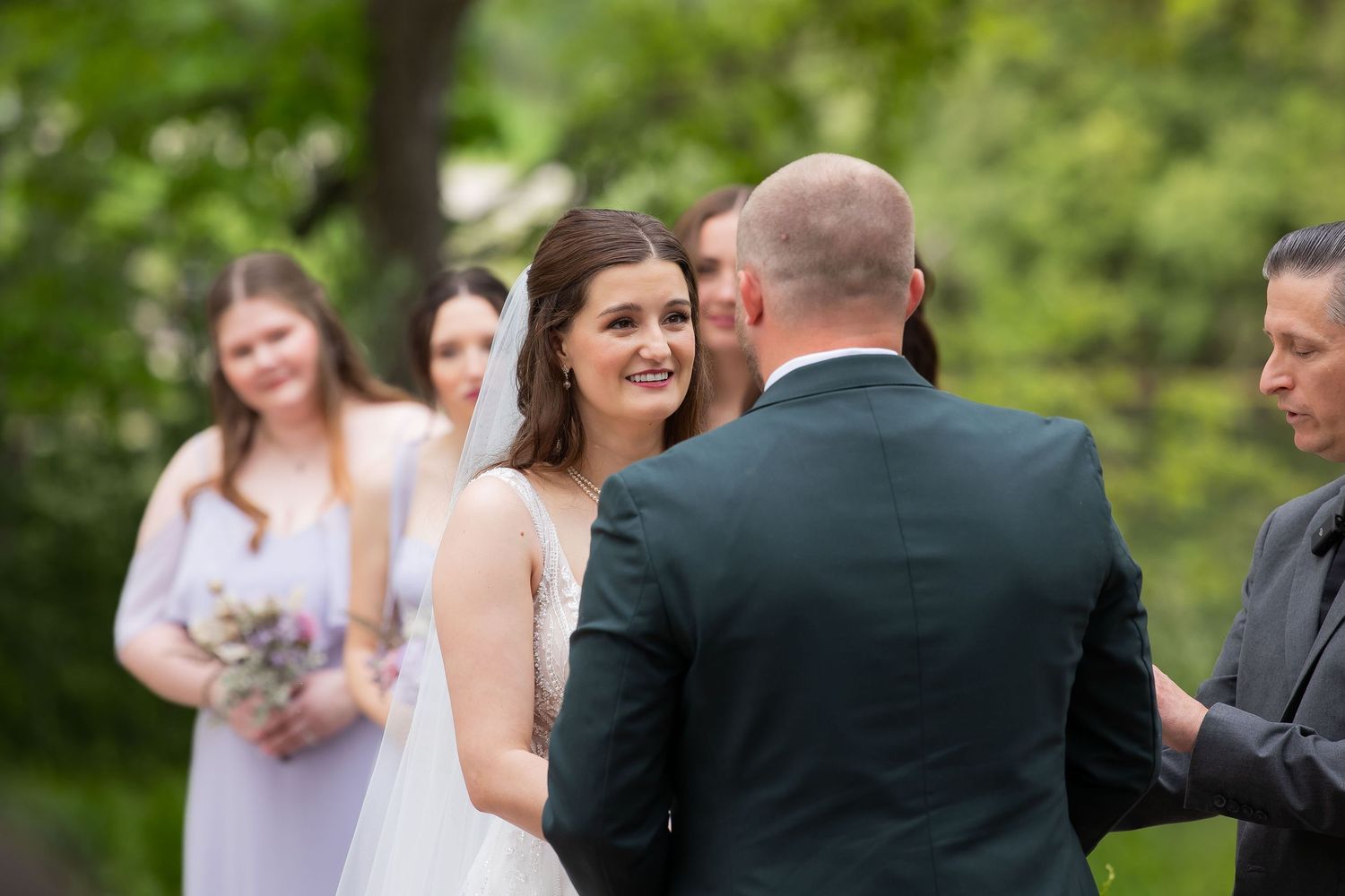 Smiling bride exchanging vows with groom during outdoor wedding ceremony with bridesmaids.