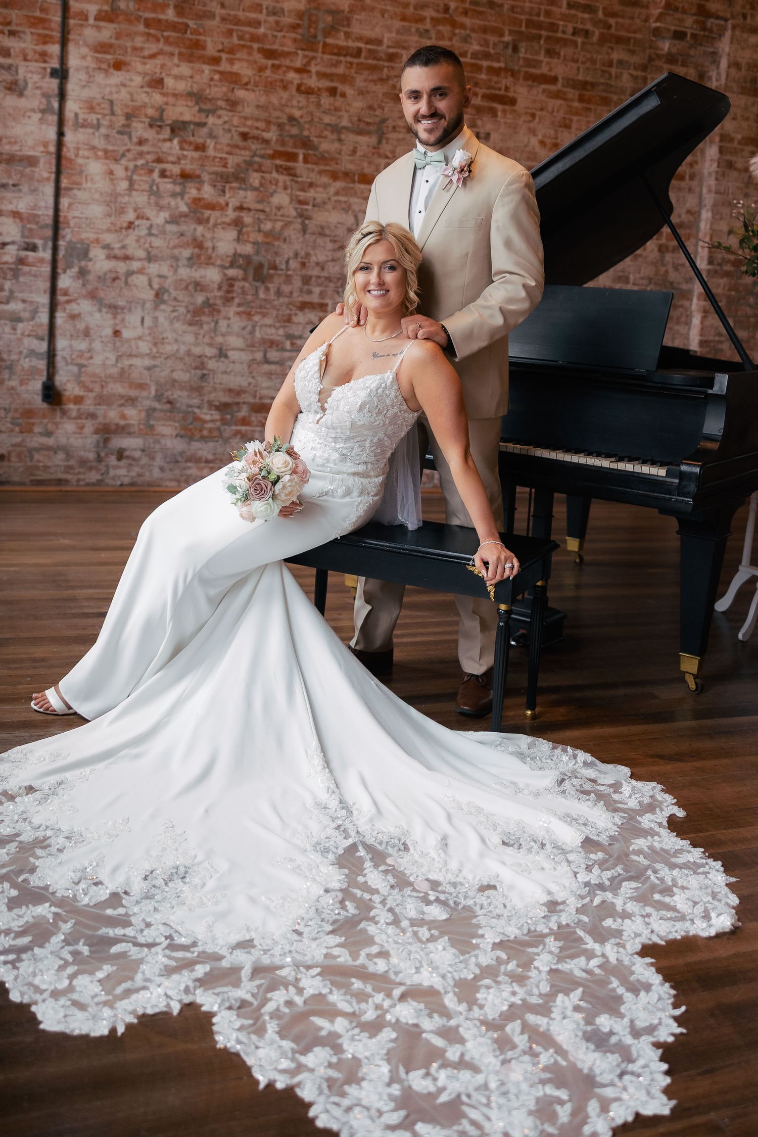 Elegant wedding photo featuring a bride in a lace train gown posing by a grand piano in an industrial brick venue.