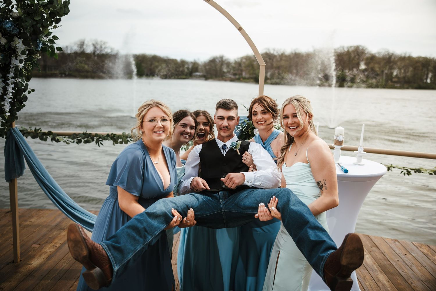Bridesmaids in teal dresses lift groom while bride laughs at outdoor lakeside wedding reception.