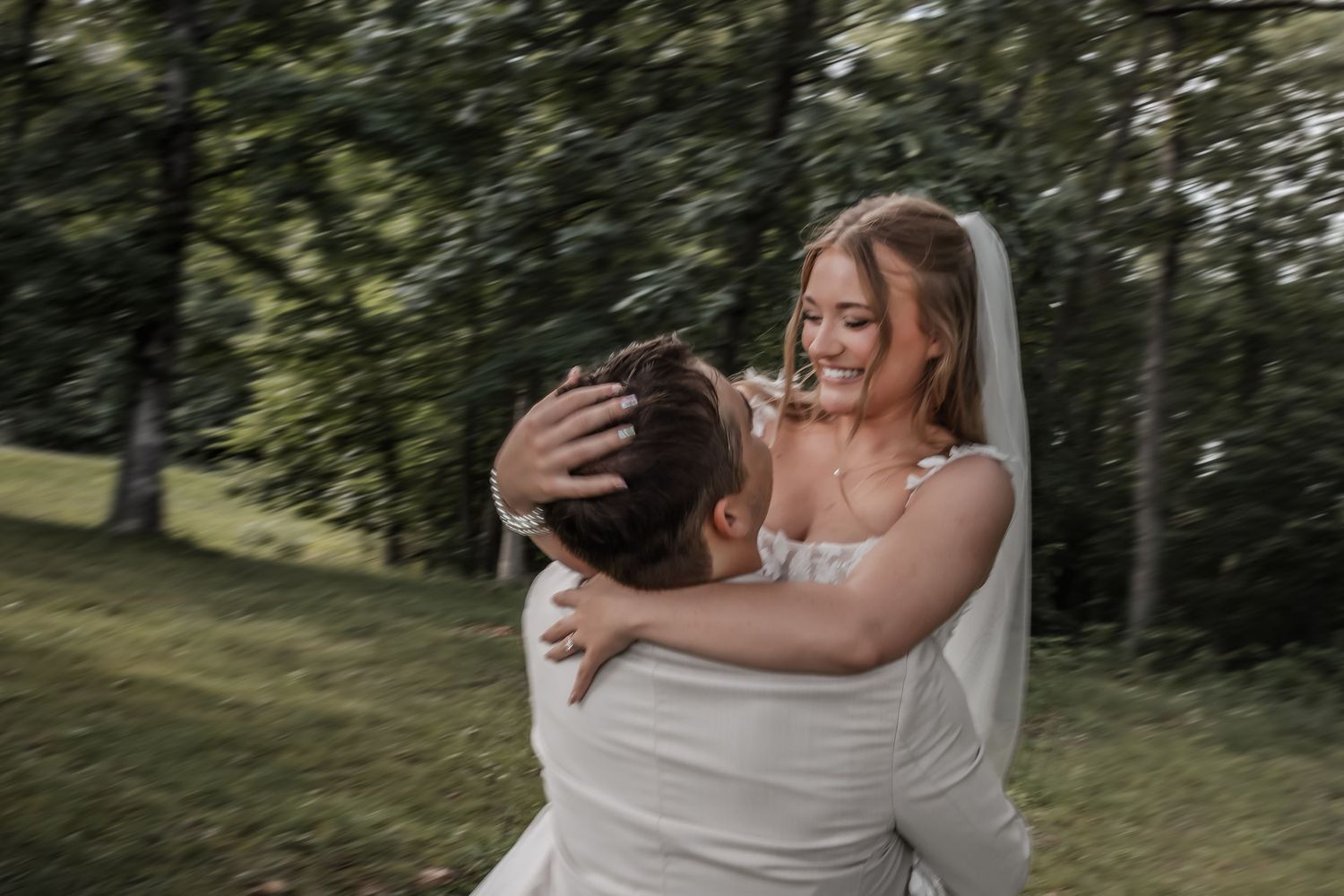 Bride in white gown and veil laughs joyfully as groom lifts her in an outdoor wedding celebration.
