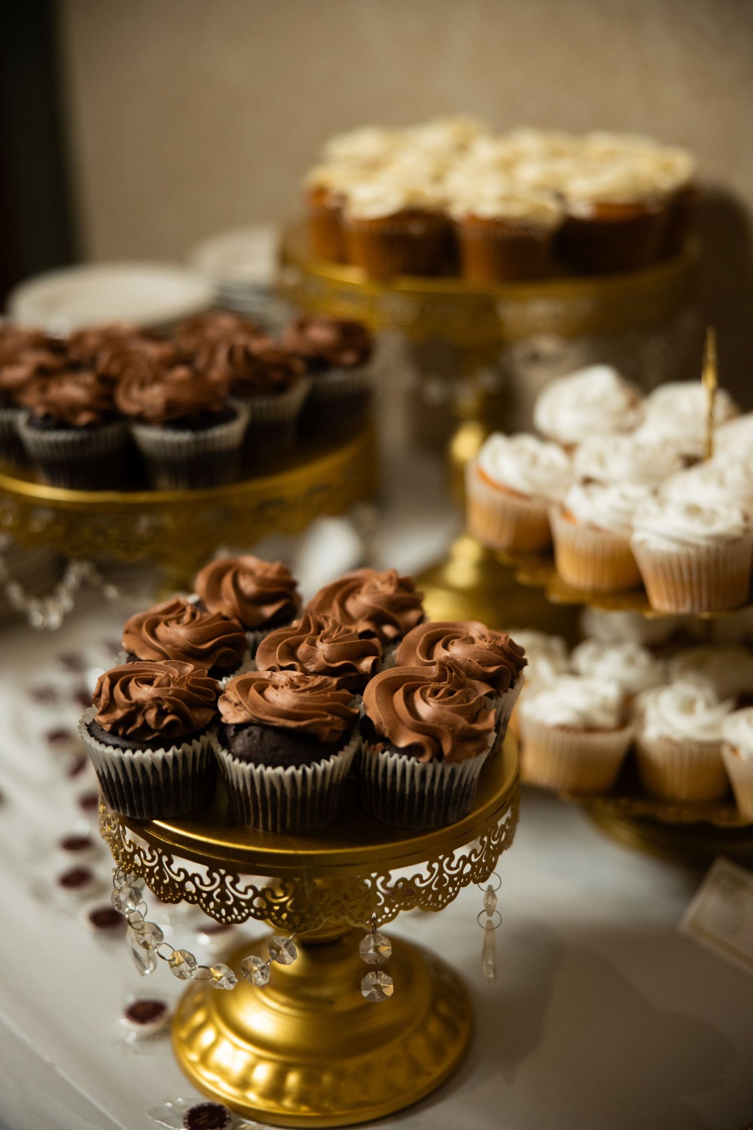 Elegant dessert table with chocolate and vanilla cupcakes on ornate gold stands at a wedding reception.