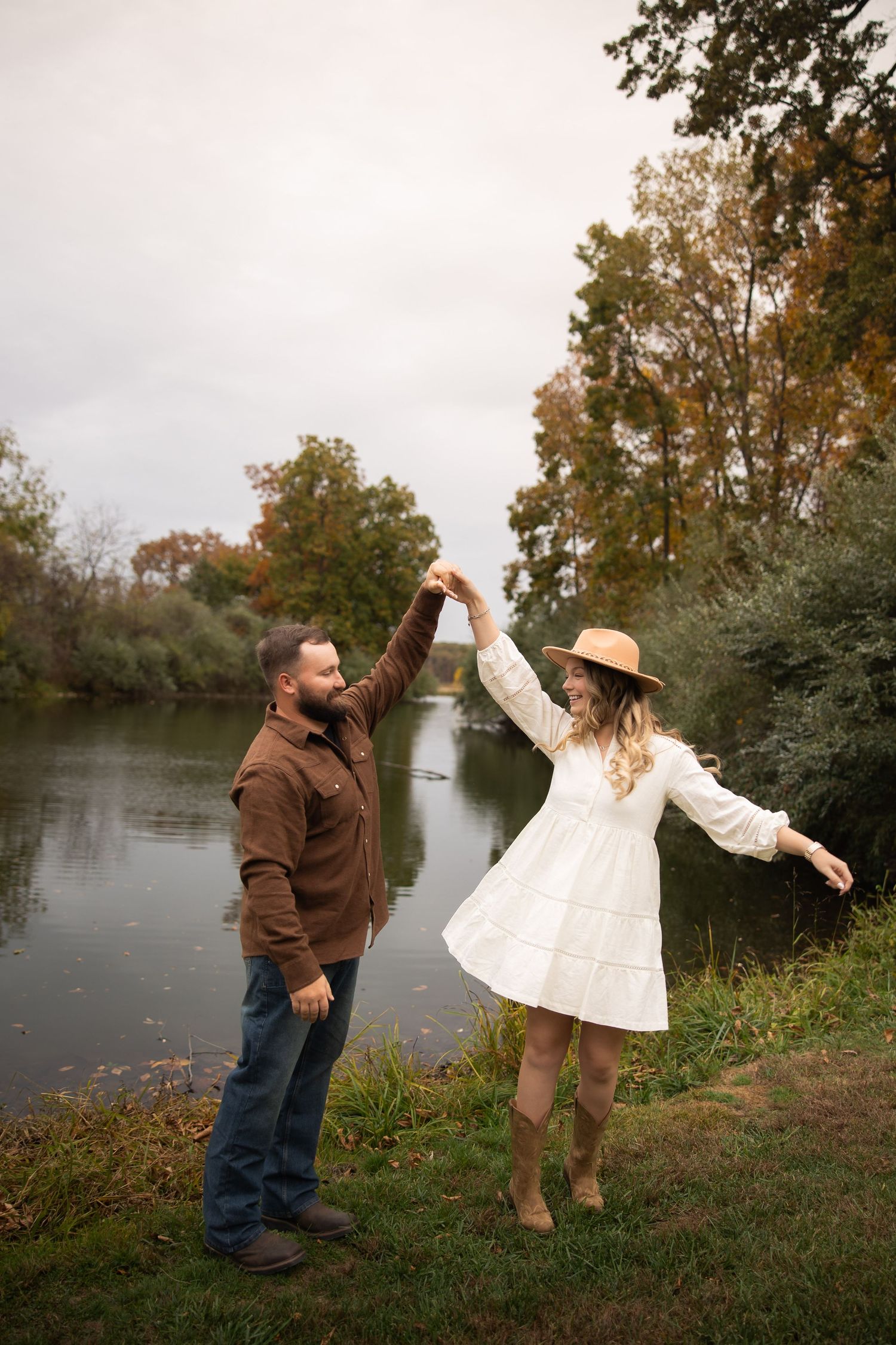 A couple dances together by a river bank during a fall photo session with trees in the background.