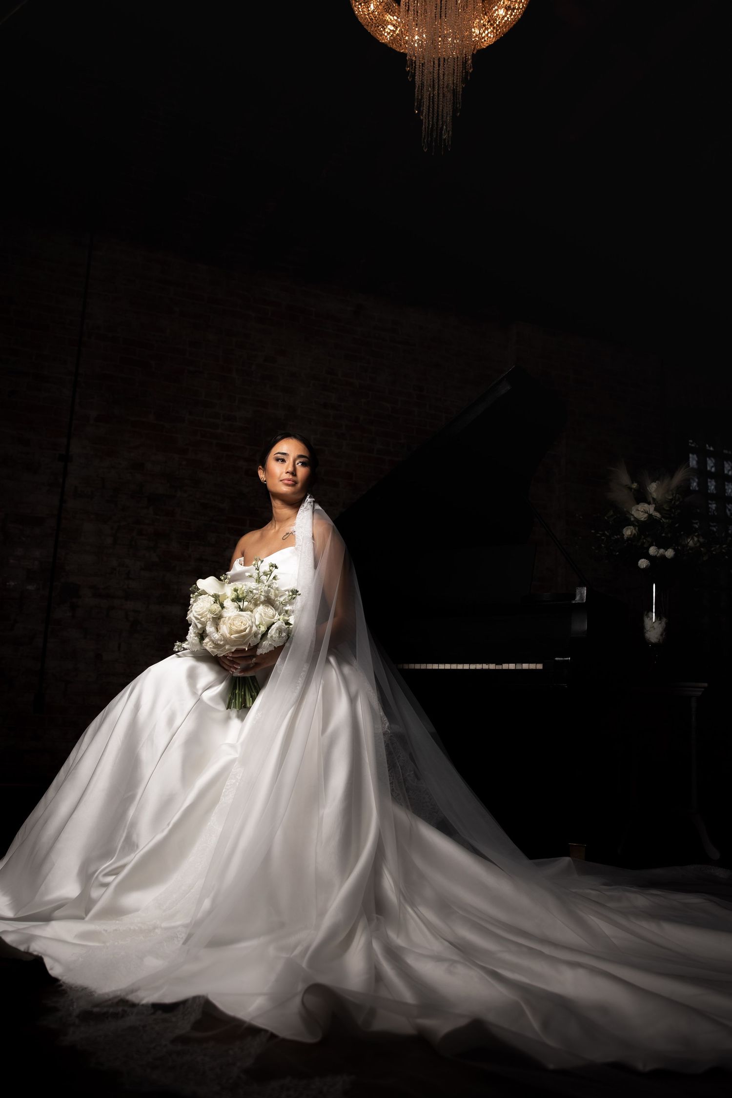 Dramatic bridal portrait with flowing white gown and bouquet under vintage chandelier in dark brick room.