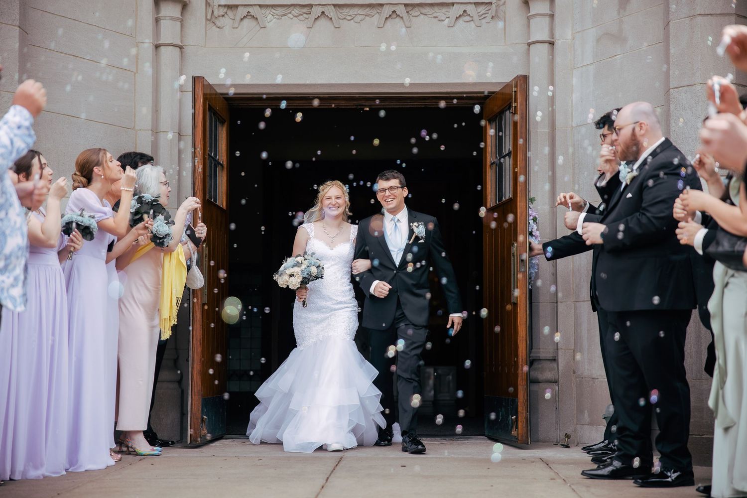 Wedding party exits church as guests throw confetti in celebration.