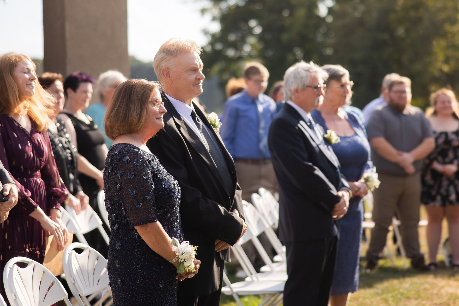 Wedding guests seated in white chairs watching a ceremony under a covered pavilion.