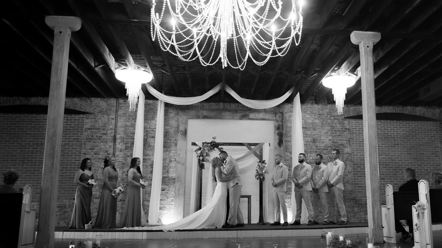 Elegant wedding ceremony under a crystal chandelier with draped fabric and white columns in a brick-walled venue.