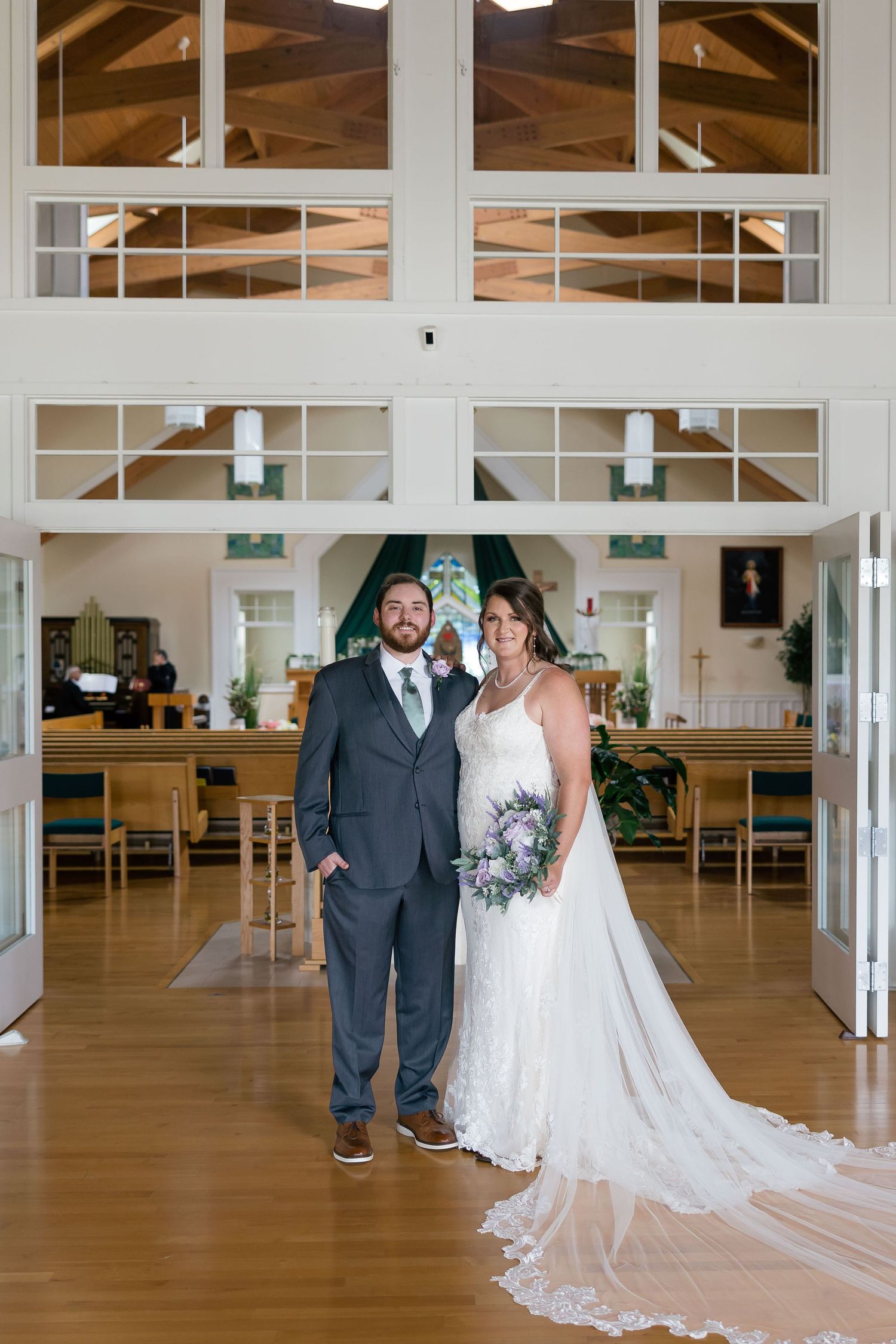 Bride and groom pose together in a church foyer, bride holding a lavender bouquet in a lace gown with a long train.