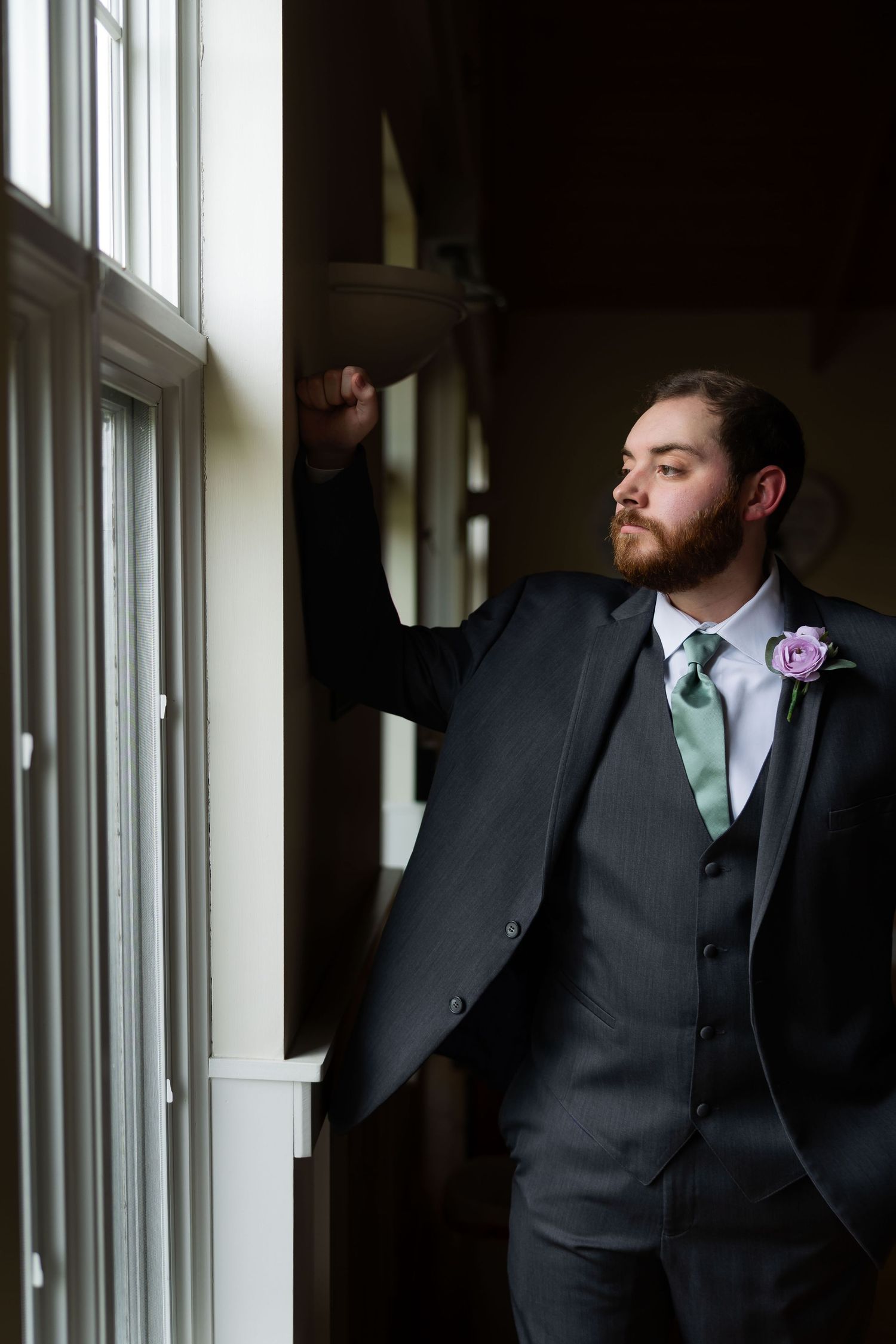 Bearded groom in gray suit with mint tie and purple boutonniere leaning against window frame.