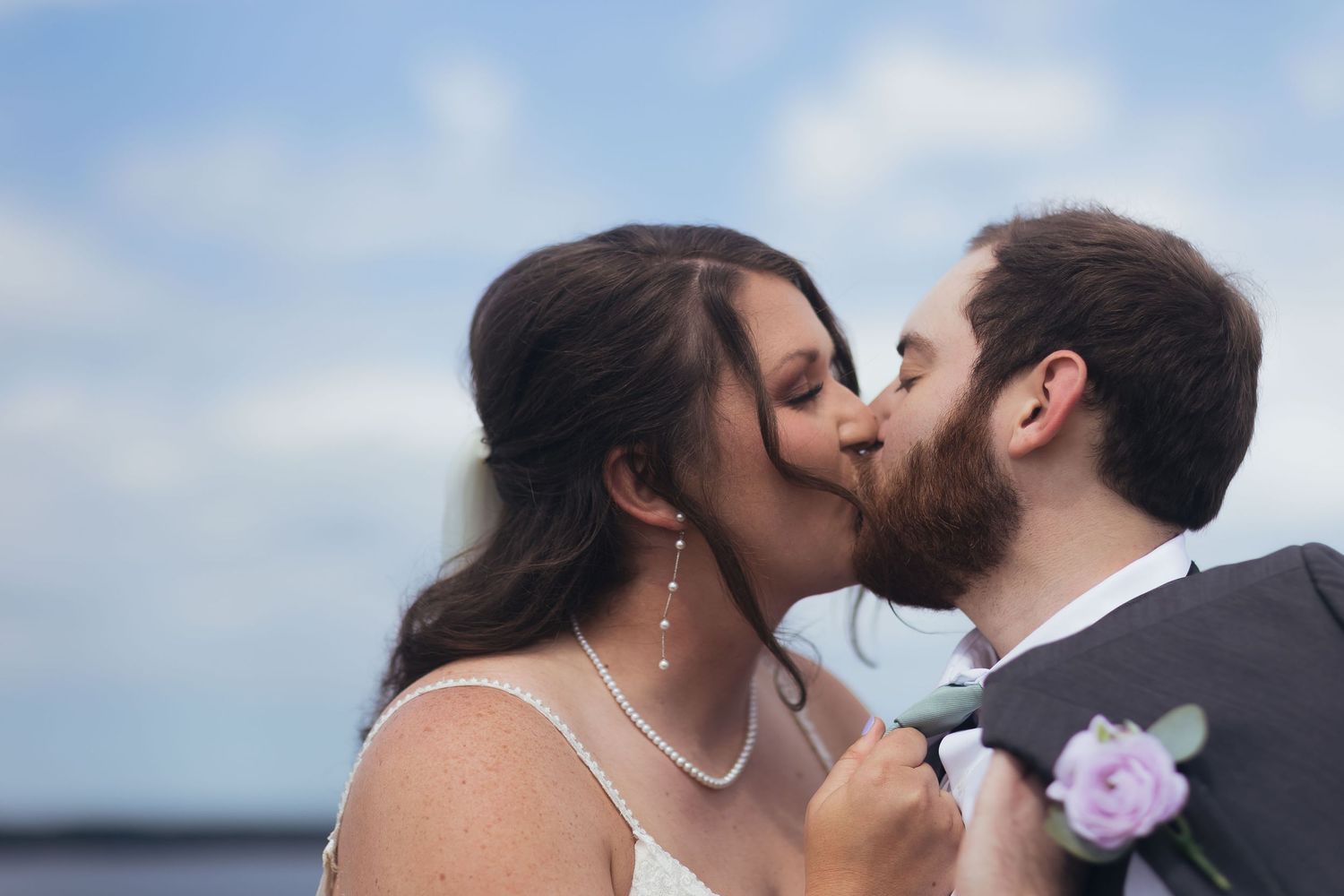 A bride and groom share a romantic first kiss outdoors on their wedding day, with a blue sky backdrop.