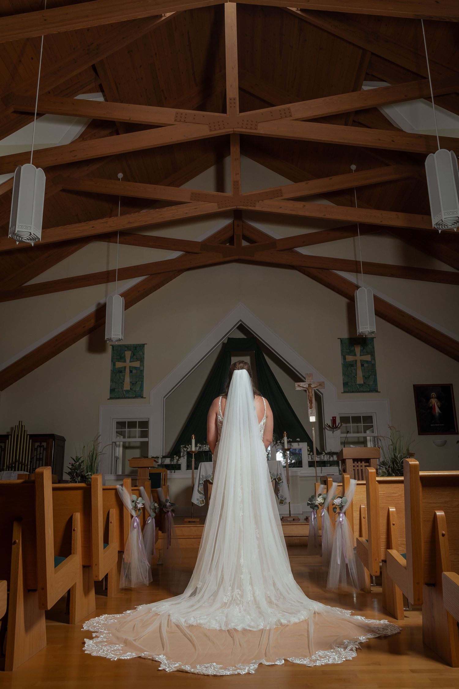 Bride in a flowing white wedding gown and long lace-trimmed veil stands at the altar of a wooden chapel.