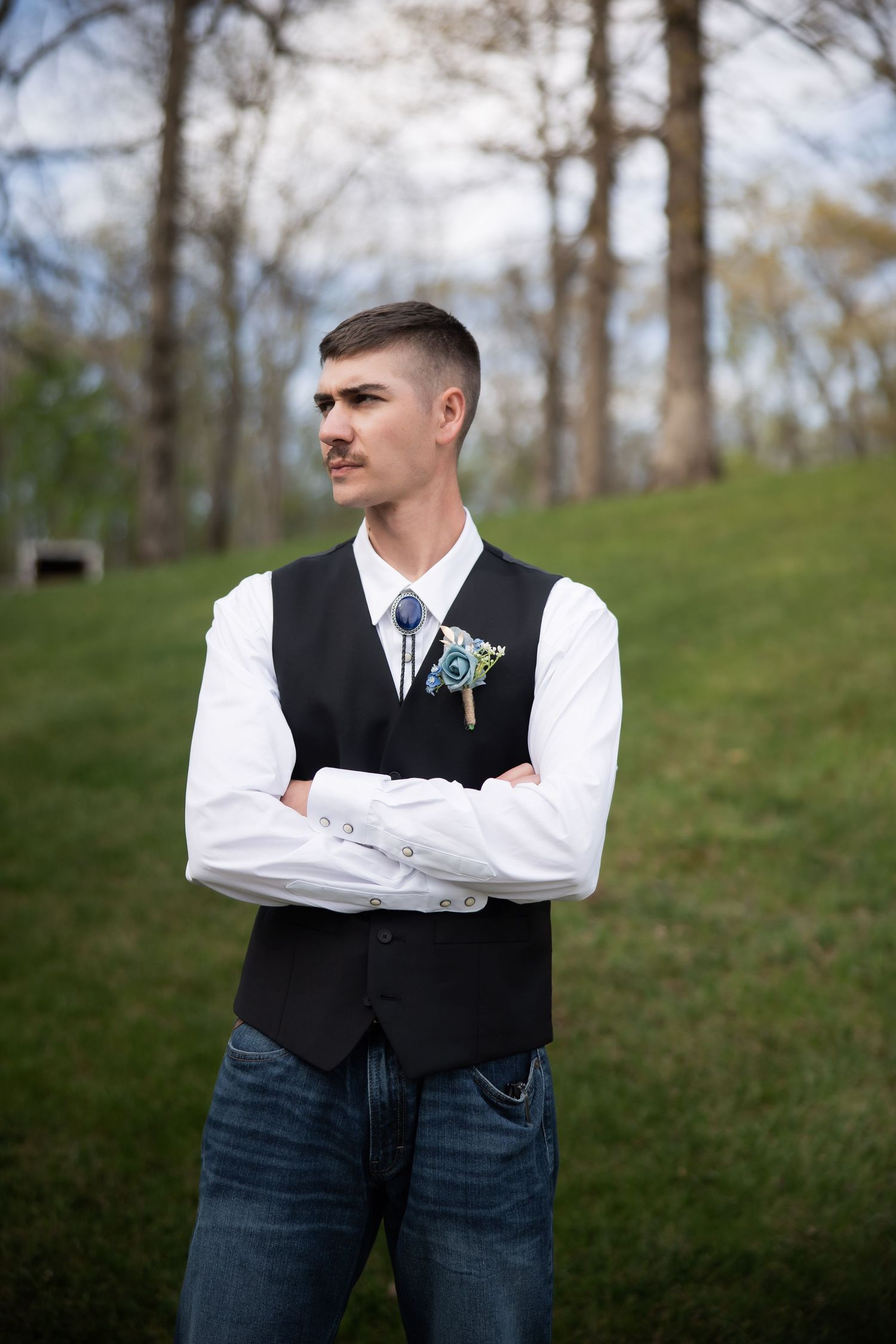 Dapper groom in black vest and jeans with boutonniere stands arms crossed outdoors at wedding.