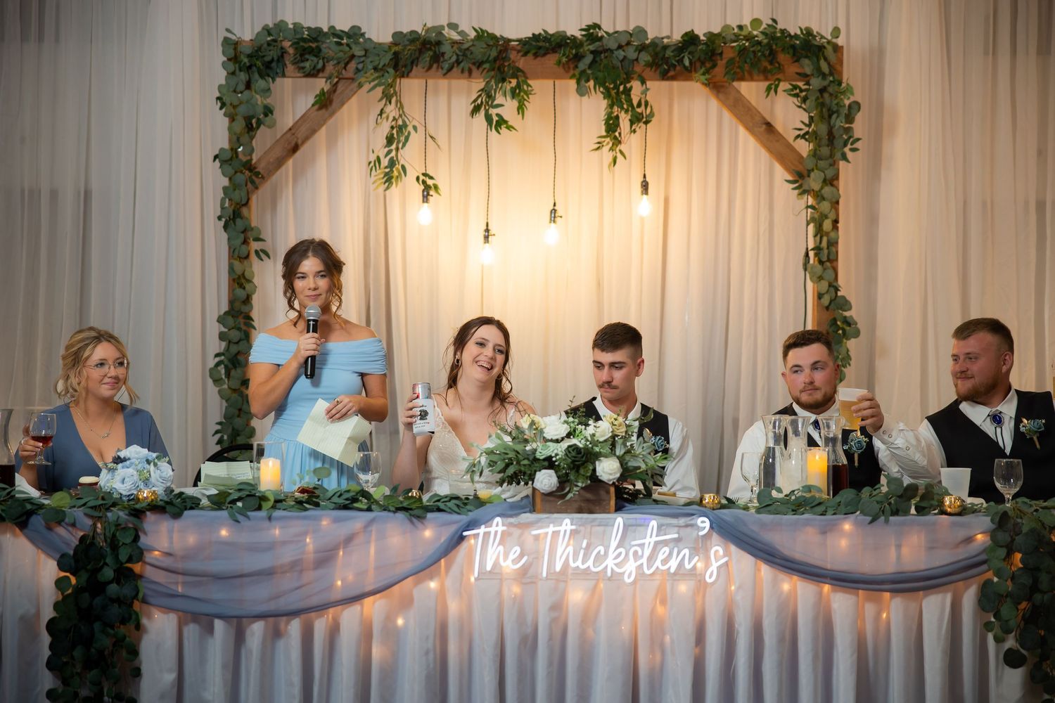 Wedding party seated at the head table as a bridesmaid gives a toast at The Thickstons reception.