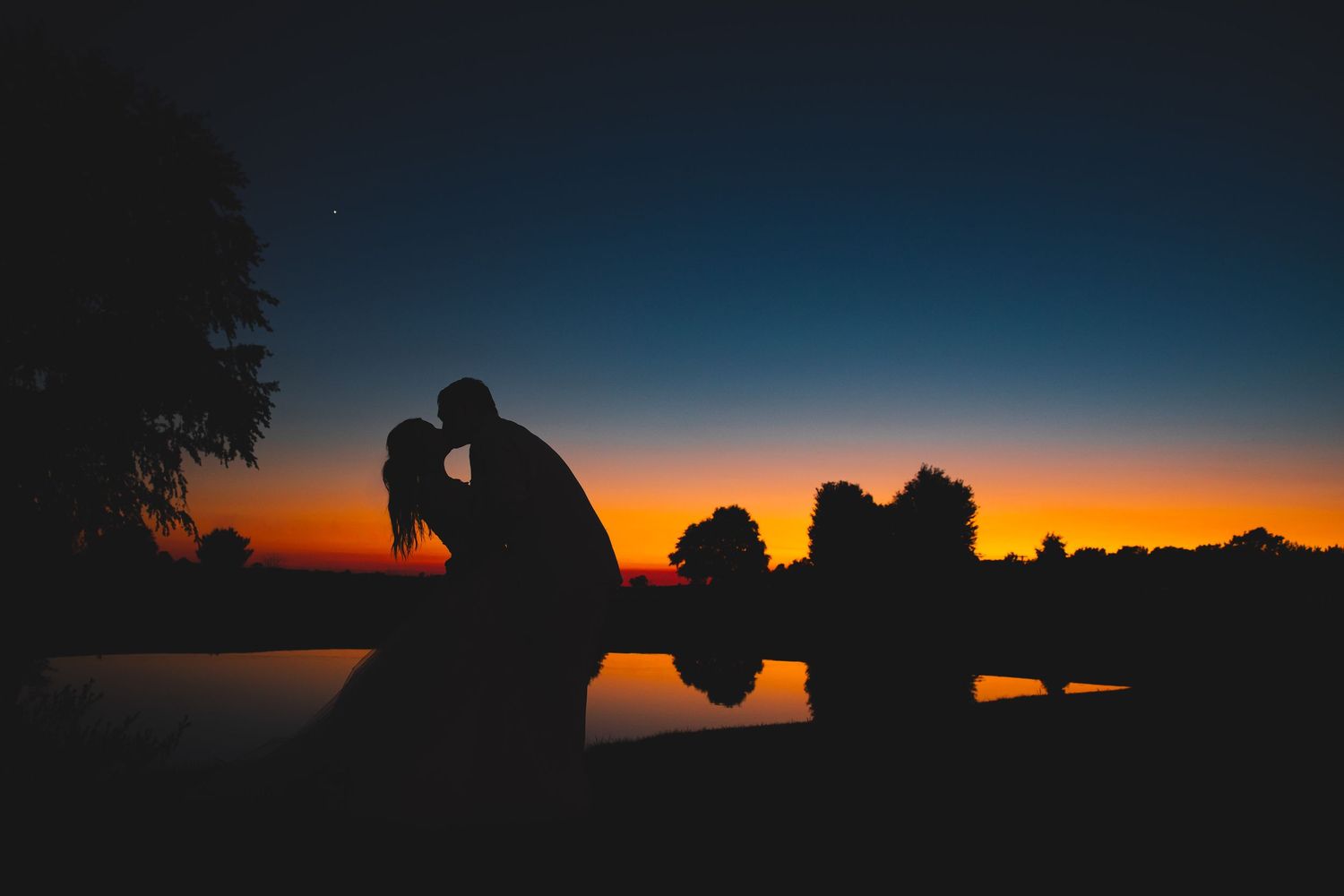 Silhouette of couple sharing a romantic kiss at sunset with vibrant orange and blue sky reflecting on water.
