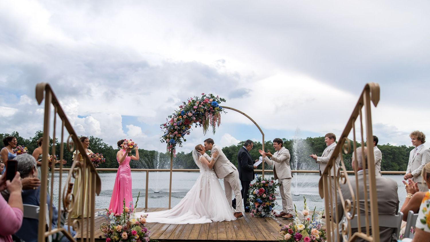 Couple kissing at outdoor waterfront wedding ceremony under floral arch on wooden dock with guests watching.
