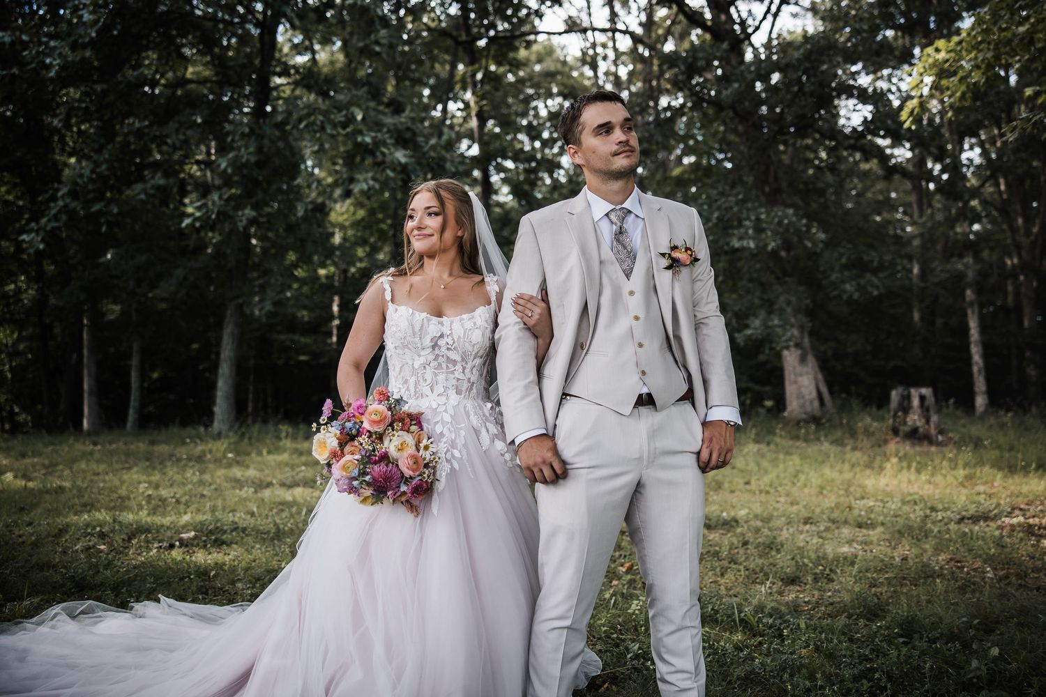 Bride and groom hold hands outdoors in lush green forest setting, bride holding colorful bouquet.