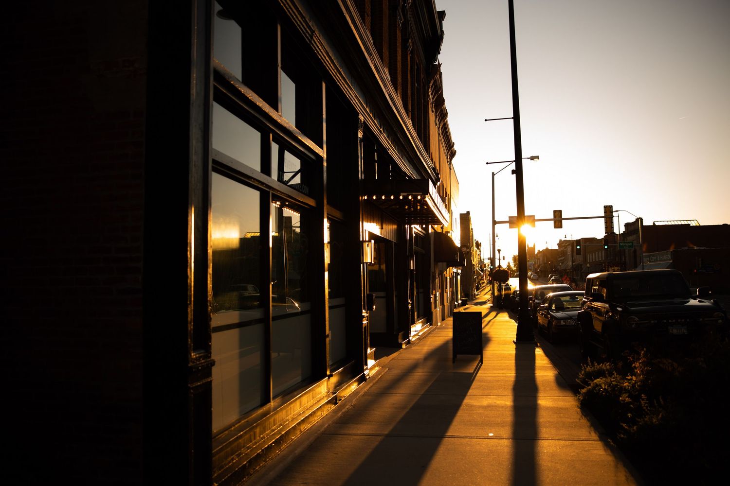 Golden sunset light illuminates a brick building storefront and empty sidewalk in a small downtown district.
