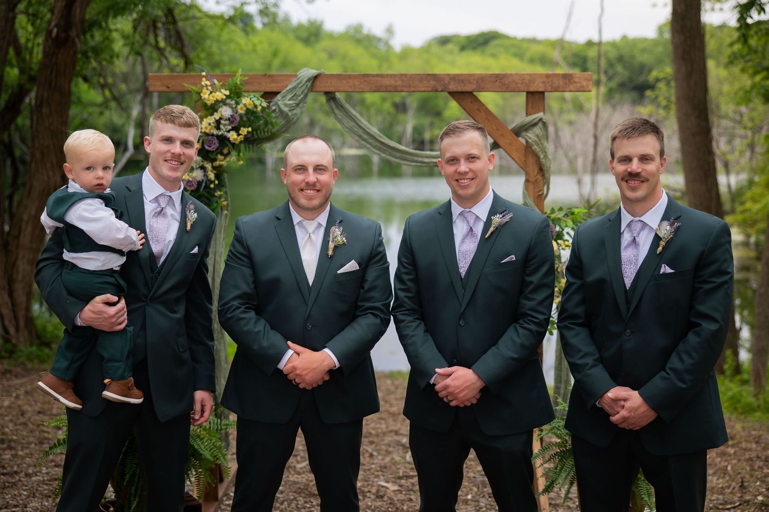 Groom and groomsmen in dark suits with lavender ties posing outdoors near wooden arch.
