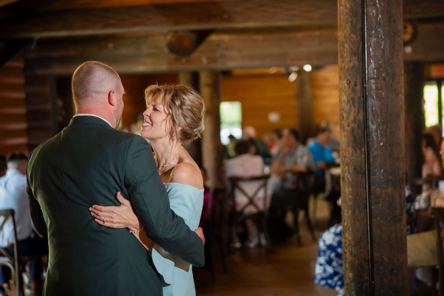 Groom and woman in light blue dress share a dance at a rustic log cabin wedding reception.