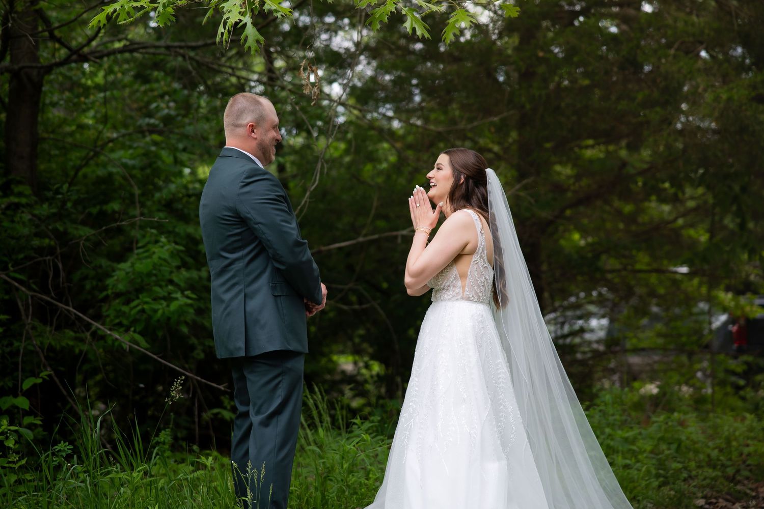 Bride in a white gown and veil reacts emotionally during a first look moment with groom outdoors.