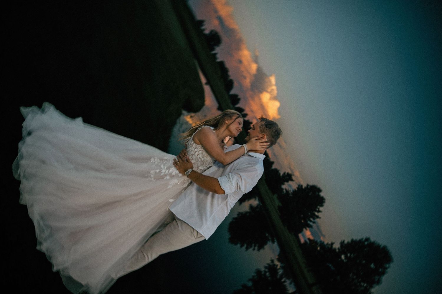Bride and groom share a romantic kiss at sunset, with dramatic sky and lush trees in the background.