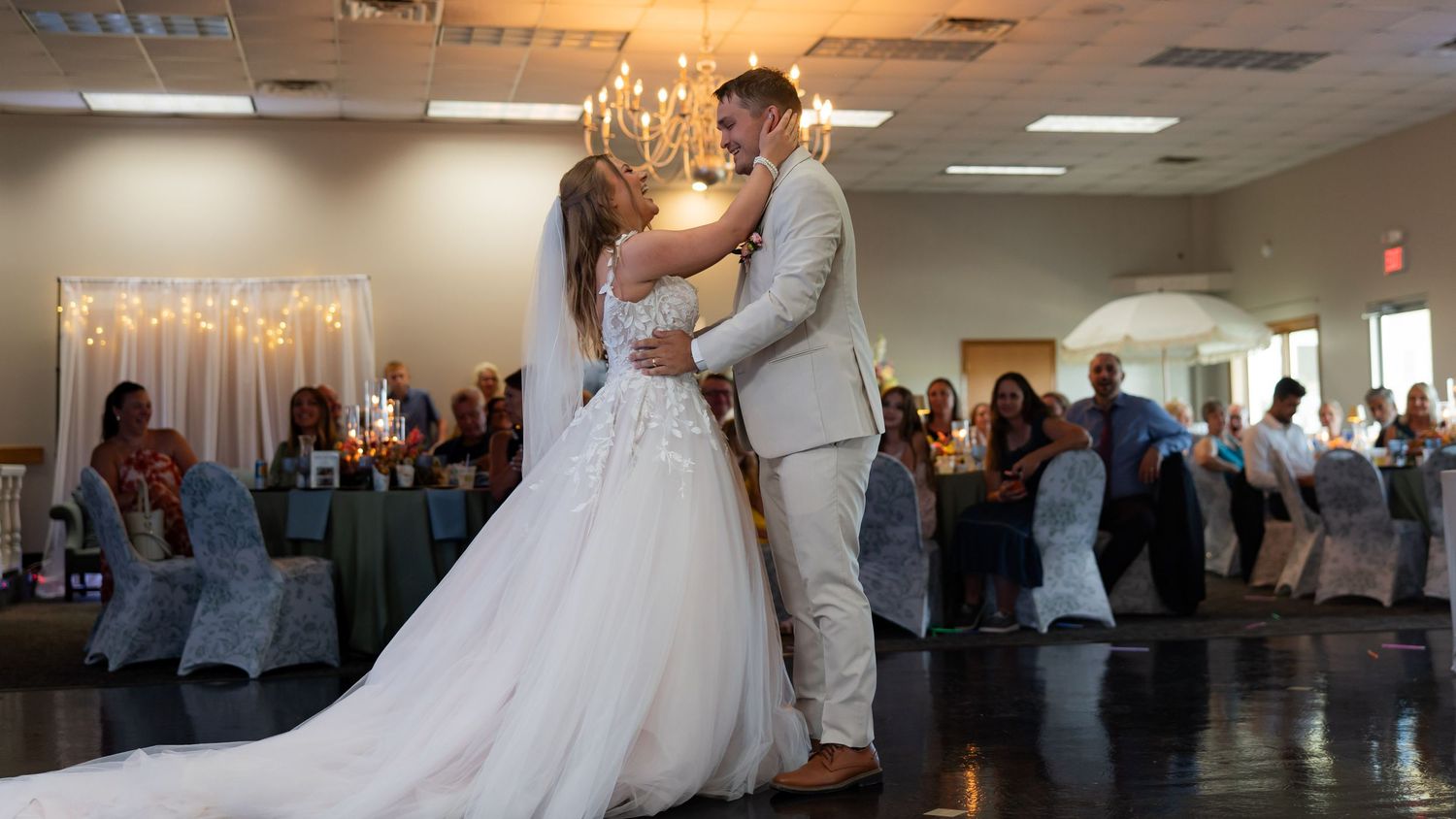 Newlyweds share romantic first dance in elegant reception hall with chandelier and guests watching.