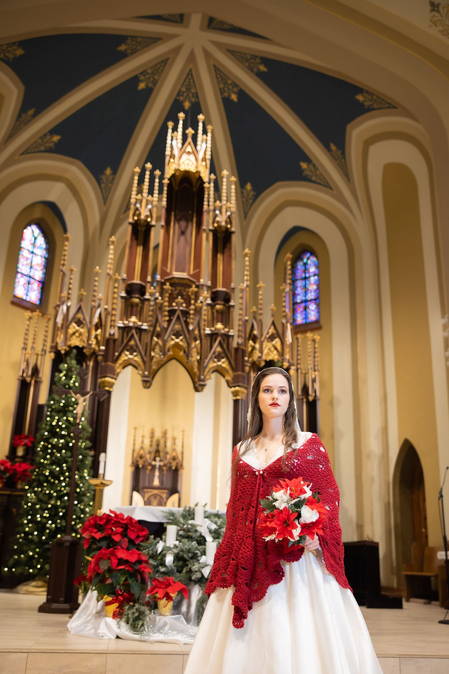 Bride in white gown and red shawl holds bouquet before ornate church altar with Christmas decorations.