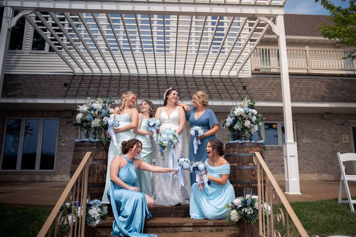 Bride in white gown surrounded by bridesmaids in blue dresses on outdoor steps with floral arrangements and pergola backdrop.