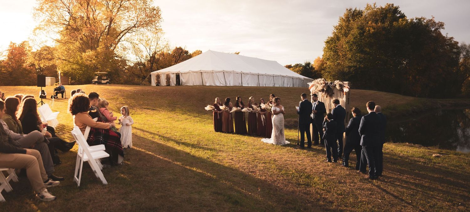 Golden sunset over an outdoor wedding ceremony with white tent and autumn trees.