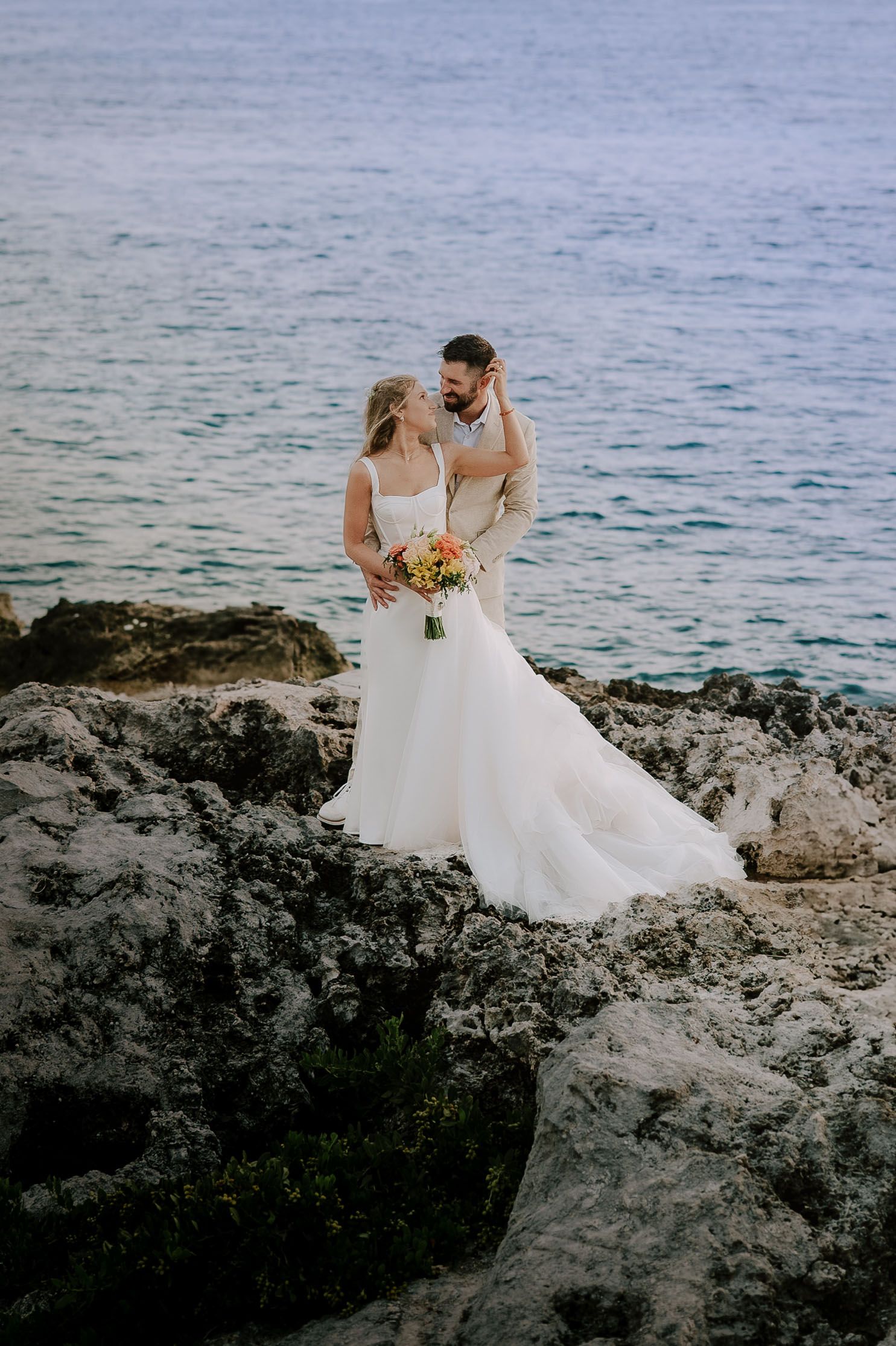 Couple in wedding attire posing on rocky oceanside cliffs during their destination wedding.