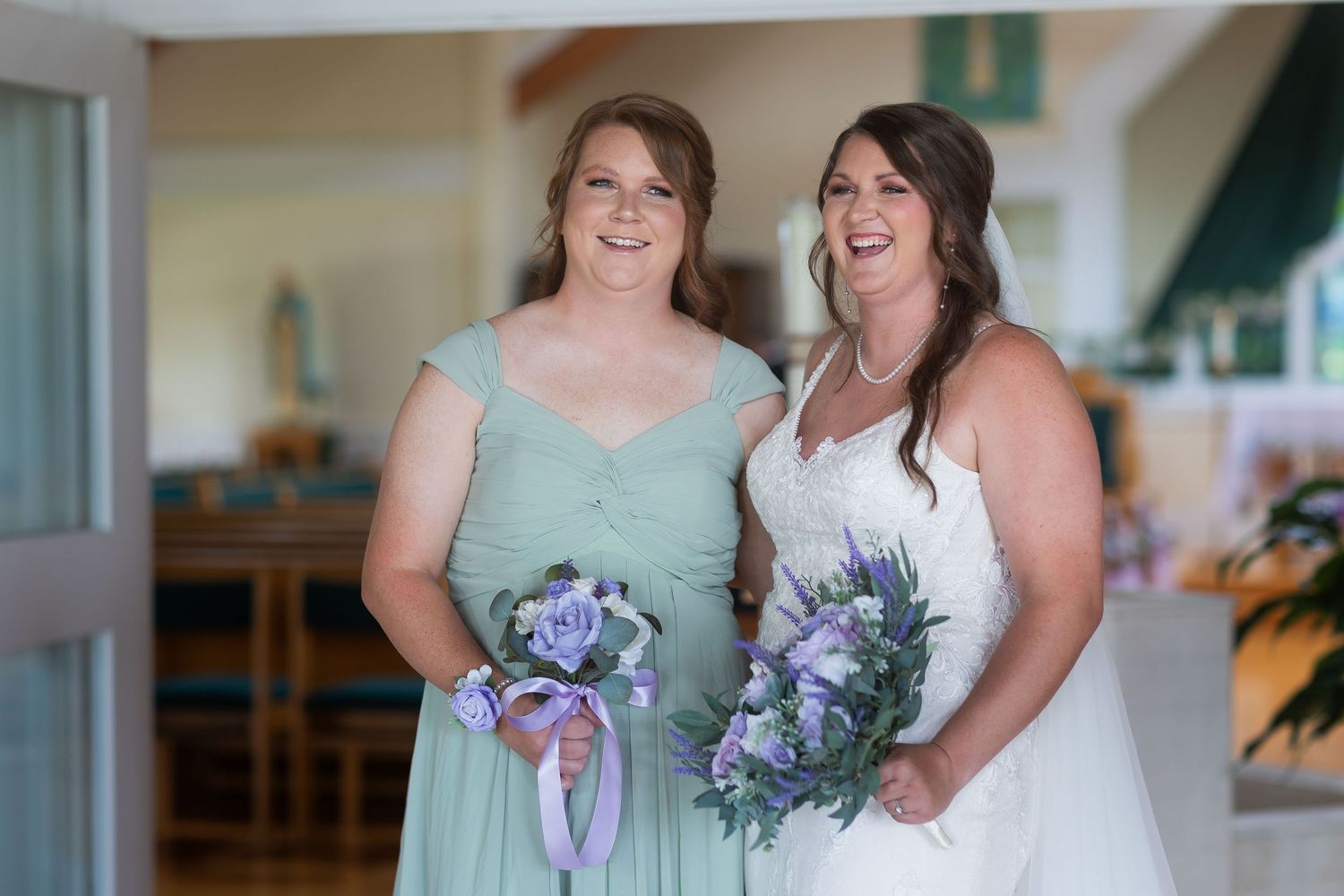 Bride in white gown and bridesmaid in mint dress holding purple bouquets smiling together indoors.