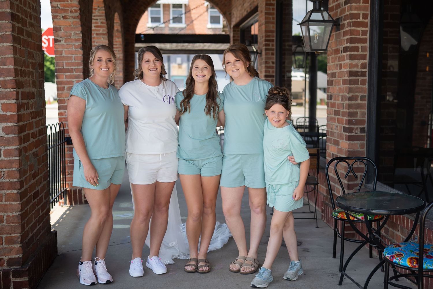 Five women in matching teal coordinated sets pose together smiling on a brick building porch for a wedding party photo.