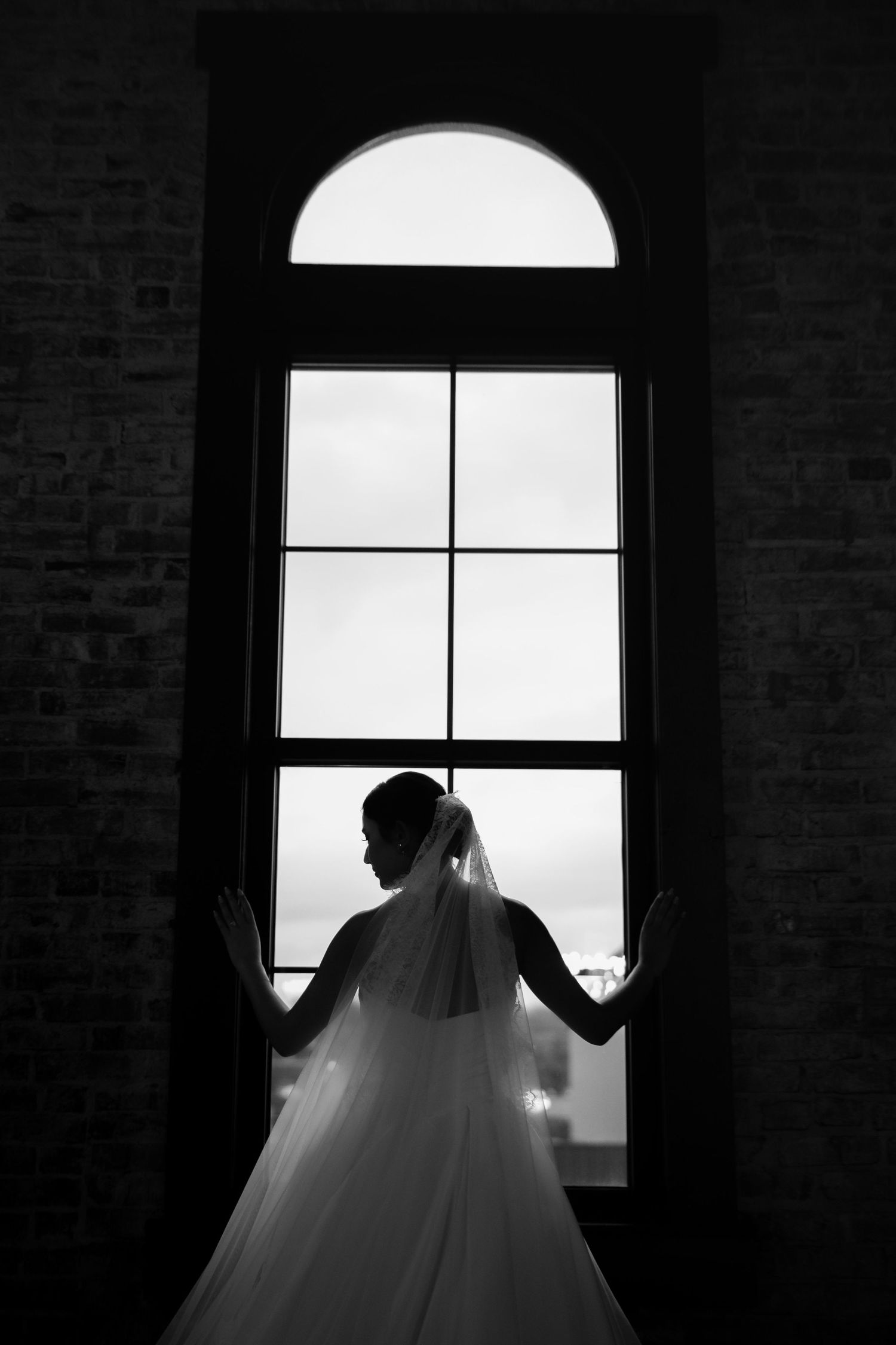 Dramatic silhouette of bride's dress against bright window in brick building.